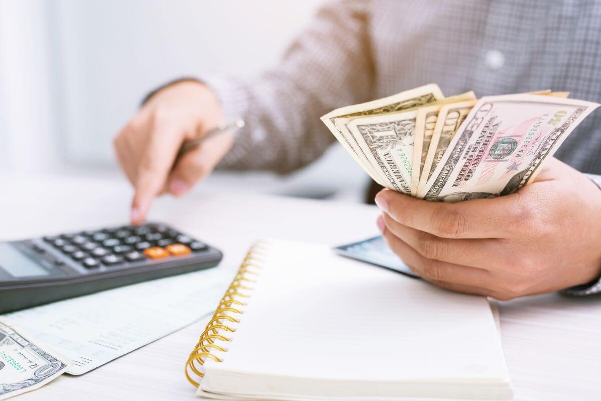 The image tells about a notebook, papers, and a man's hand holding dollar bills while calculating referring to how reevaluating your long-term financial goals and aspirations helps you focus on what truly matters.