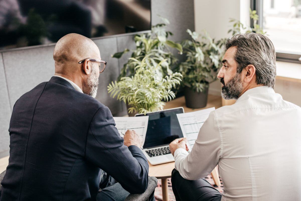 The photo tells about a laptop on the table, plants, and an advisor explaining the paper to his client which means to consult with a wealth manager that help you ensures you make smart decisions for the future.