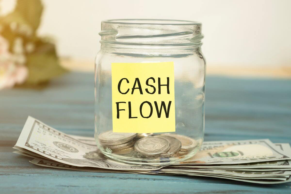 The photo tells about dollar bills on the table, a jar with coins, and a note indicating to monitor and optimize cash flow to ensure you're living comfortably while staying financially secure for the long haul.
