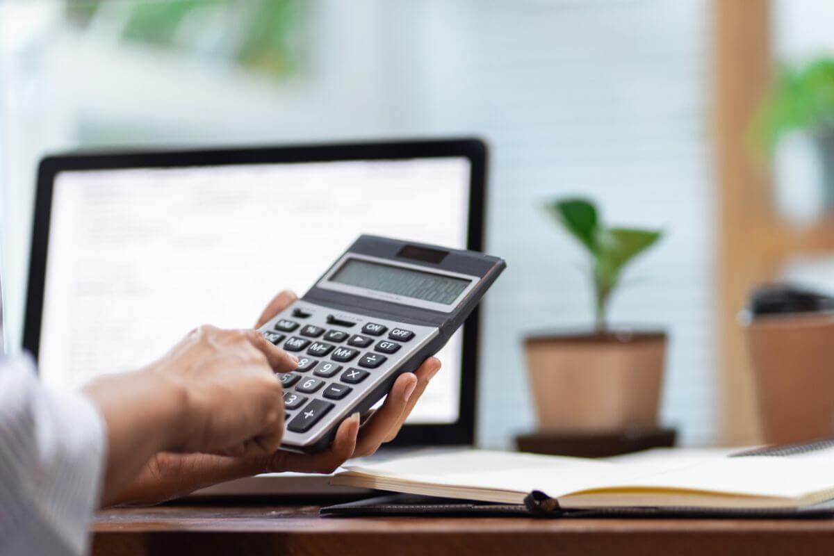 The photo of a plant on the table, a notebook, a laptop, and a man's hand calculating indicates to regularly reassess your budget to make sure your spending aligns with your long-term goals and financial independence.