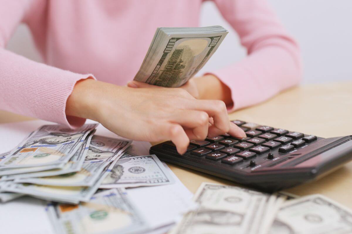 The photo of dollar bills on the table and a girl's hand calculating while holding dollar bills means to focus on enjoying financial freedom responsibly but keep your focus on long-term financial security and growth.