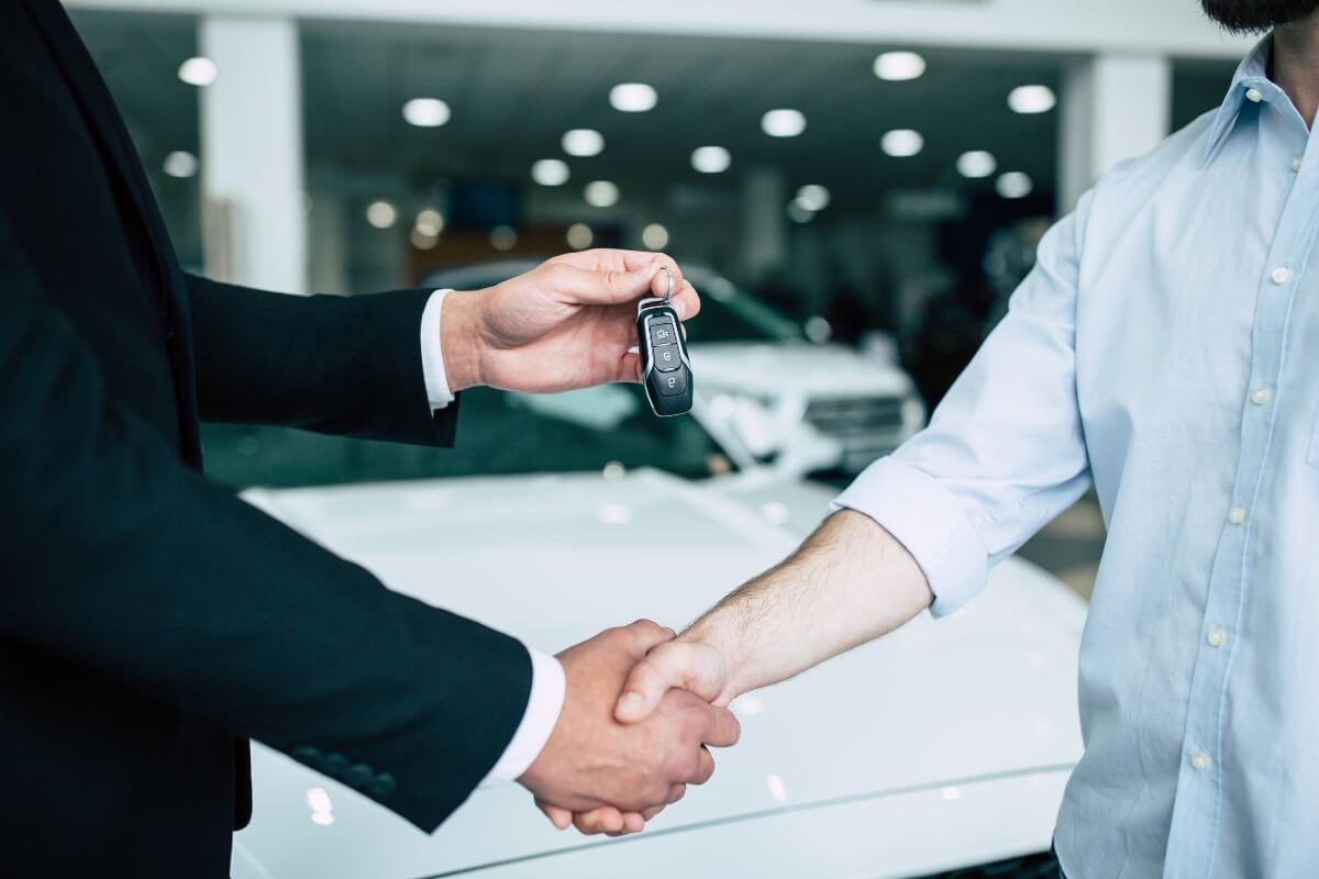 The photo shows car behind, a man in suit giving the car key to the customer while shaking hands together referring to avoid large purchases and keeping expenses low lets you save more each month.