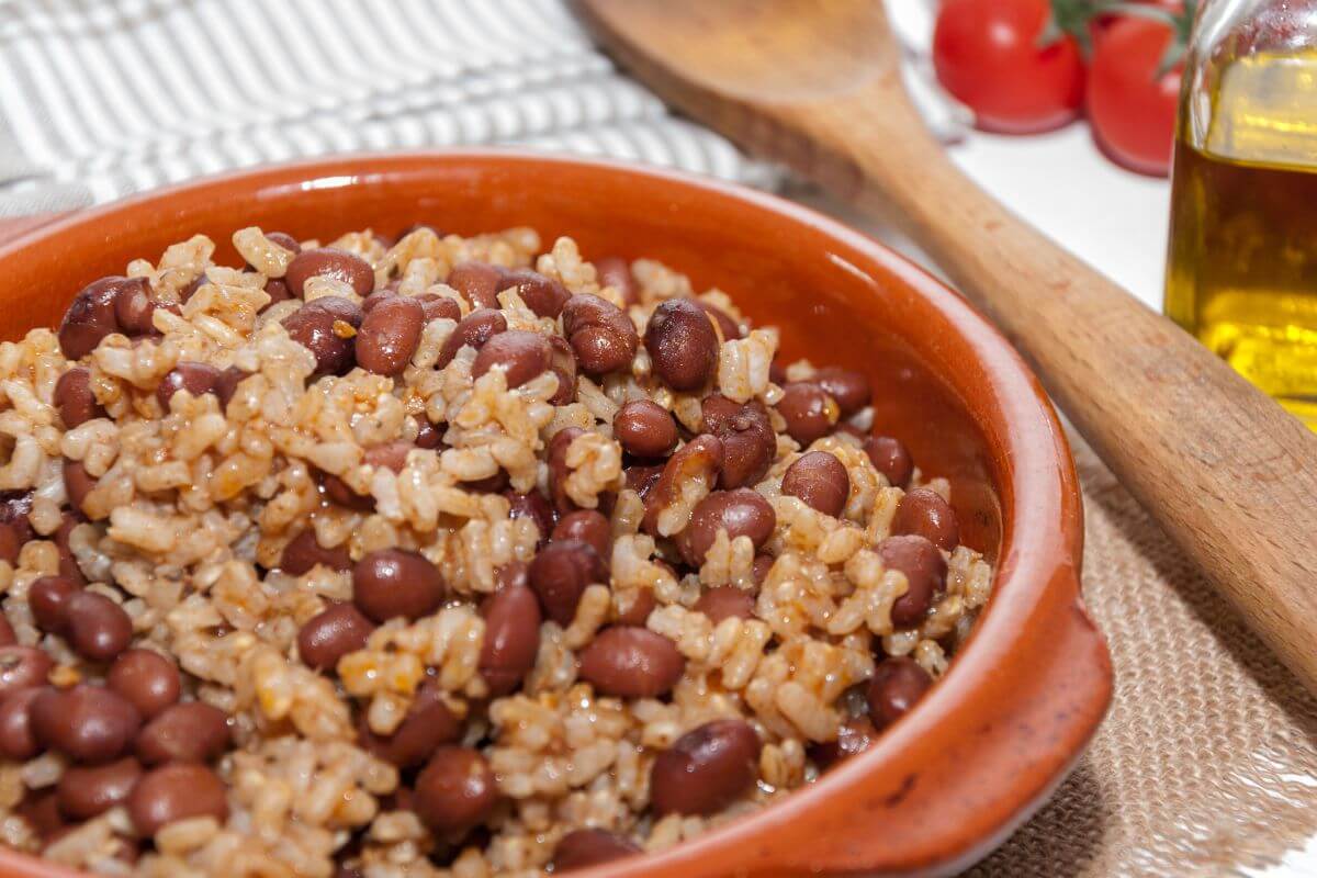The image shows a wooden spoon, tomatoes, oil in a glass container, and a wooden plate filled with rice and beans which is a budget-friendly meal perfect for stretching your dollar while still eating well.