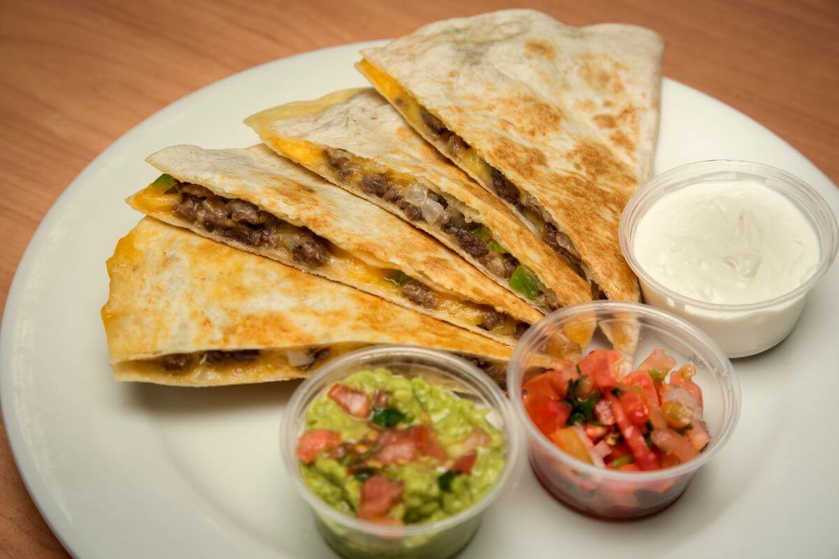 The photo tells about quesadillas with guacamole and sour cream on the plate which are quick to make and perfect for saving on food.