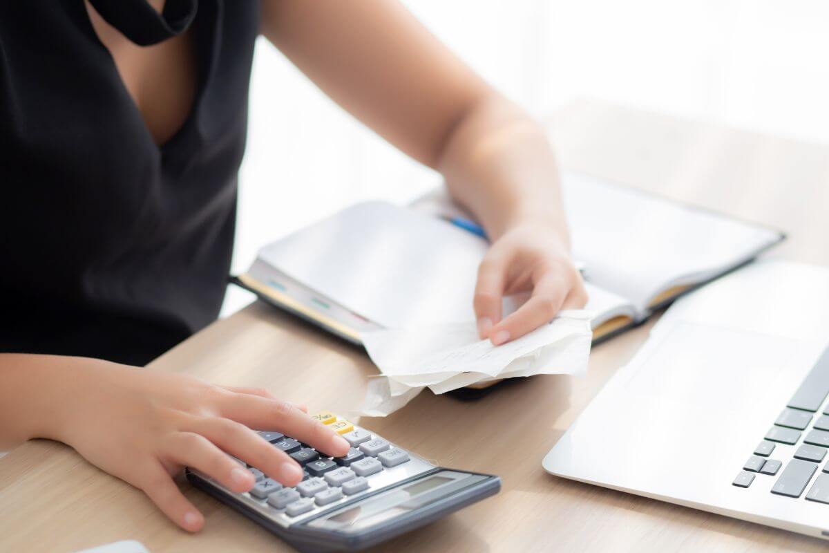 The photo shows a laptop, a notebook, and a girl calculating the papers on her hand which means to downsize expenses to free up money for savings or paying off debt.