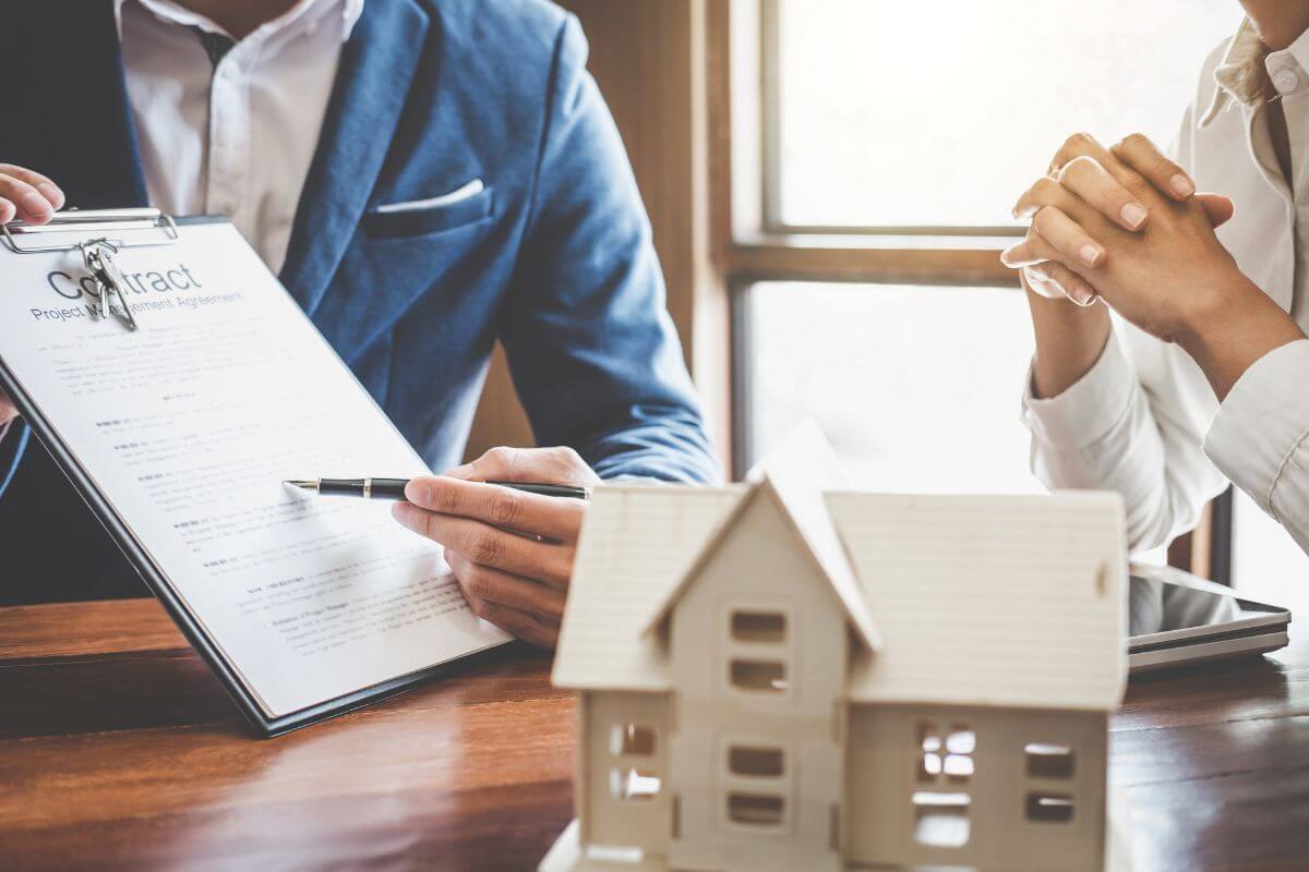The picture shows a mini figured house, a man in a suit explaining the clipboard with a contract, and a client listening to him which means owning and managing real estate properties creates wealth through consistent rent and increasing property values.