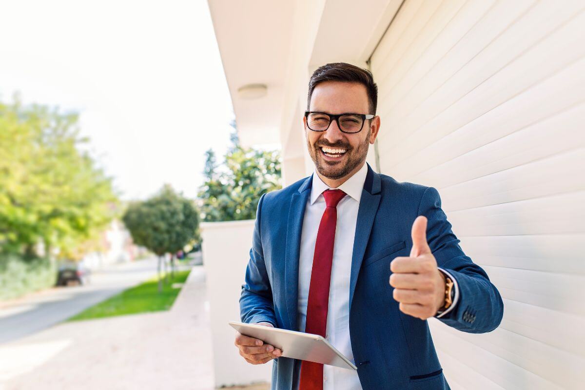 The phot tells about a man in a suit holding a tablet and doing a thumbs-up sign referring to starting and scaling a successful business as a proven path to financial independence and long-term success.