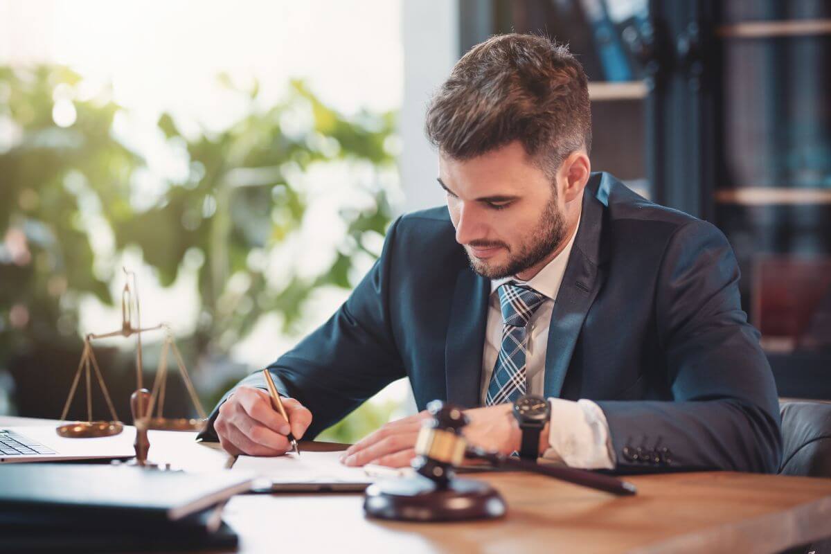 The photo tells about a laptop, a balanced weighing thing, a gavel, and a man in a suit writing in the notebook which refers to high-paying professional roles contributing heavily to financial success and long-term wealth.