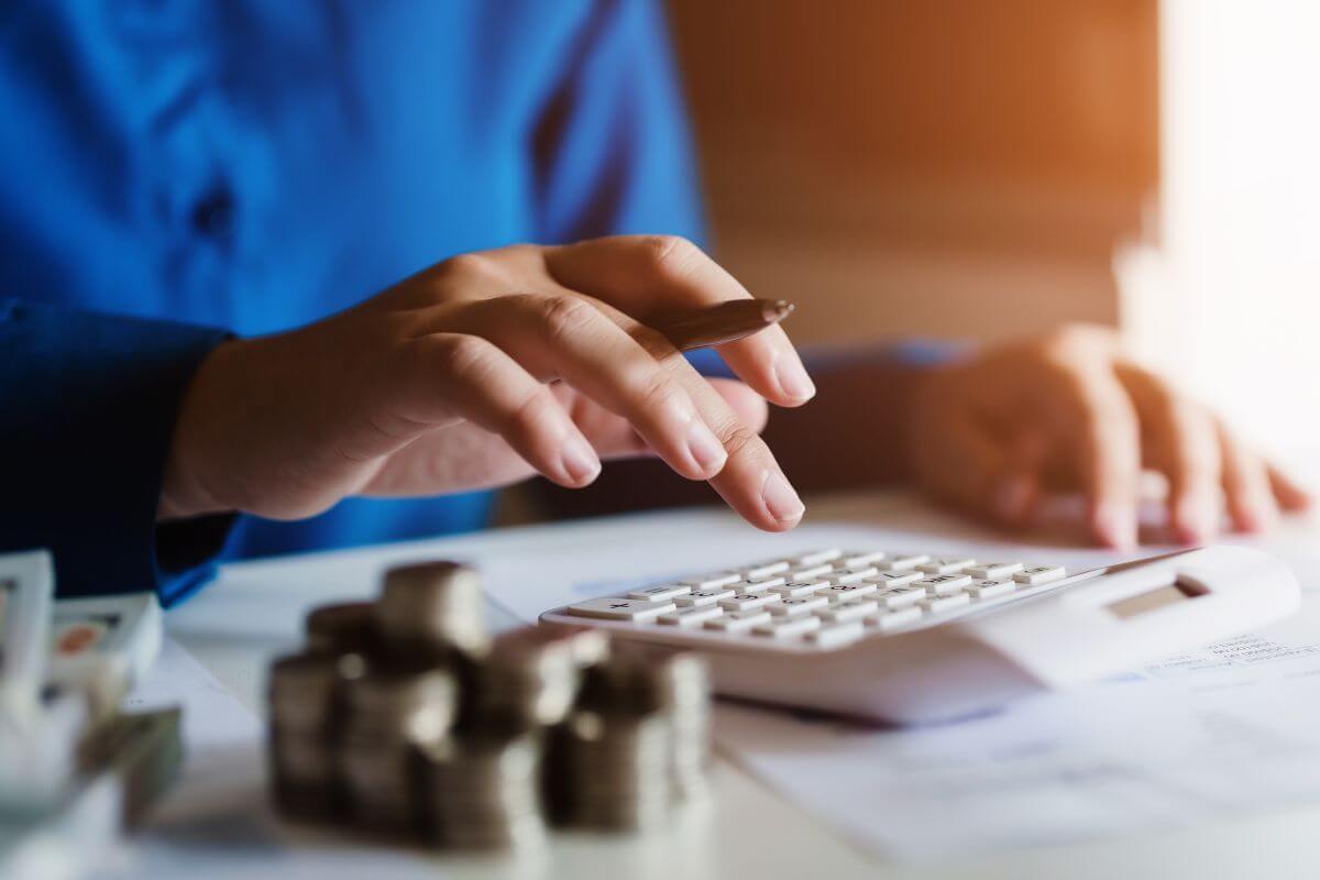 The image shows coin stocks, dollar bills, and a girl's hand calculating while checking on the paper which means not tracking all expenses can ruin your budget and leaving you with less  money than you think.