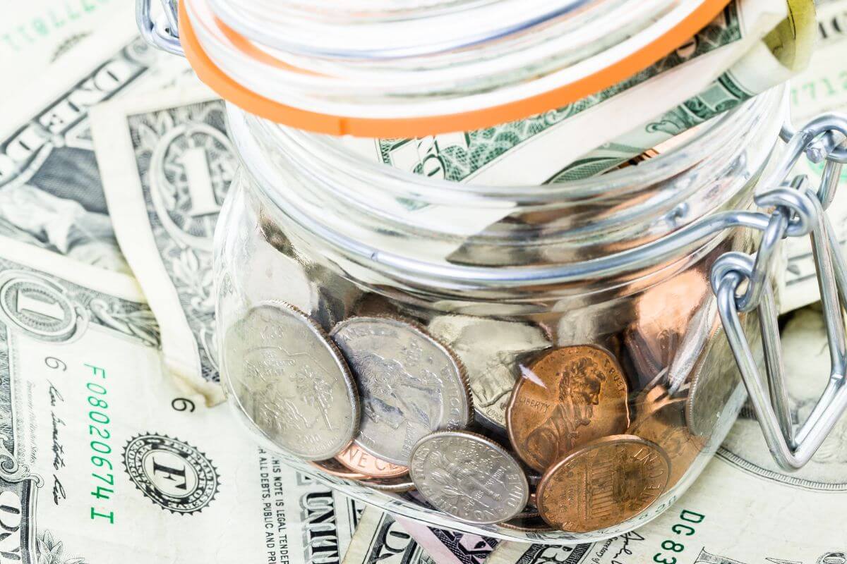 The picture tells about dollar bills on the table and a jar filled with coins and dollar bills indicating to forgetting to include irregular costs can crush your budget and planning for them ensures you don't have to dip into savings.