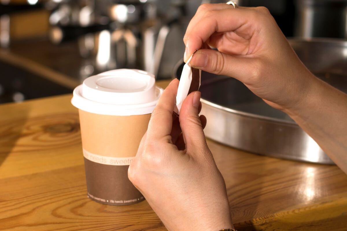 The image tells about a girl's hand tearing the sugar to add in her coffee in a cup to go referring to ignoring daily small purchases that eat away at your budget and can add up to hundreds of dollars.