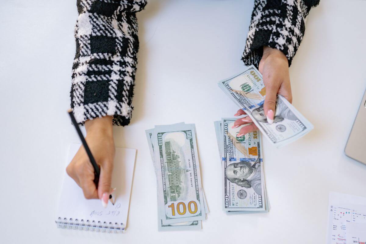 The picture shows money grouped into two on the table and a girl's hand holding dollar bills while writing in the mini notebook which refers to setting unrealistic spending limits is a quick way to set up for failure and end up overspending later.