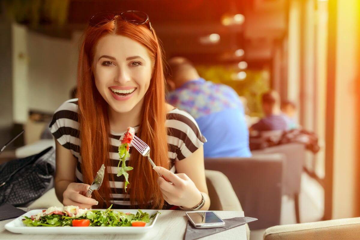 The picture tells about a girl eating at a restaurant indicating that overspending on non-essentials can add to your expenses quickly, finding cheaper alternatives can also help save money.