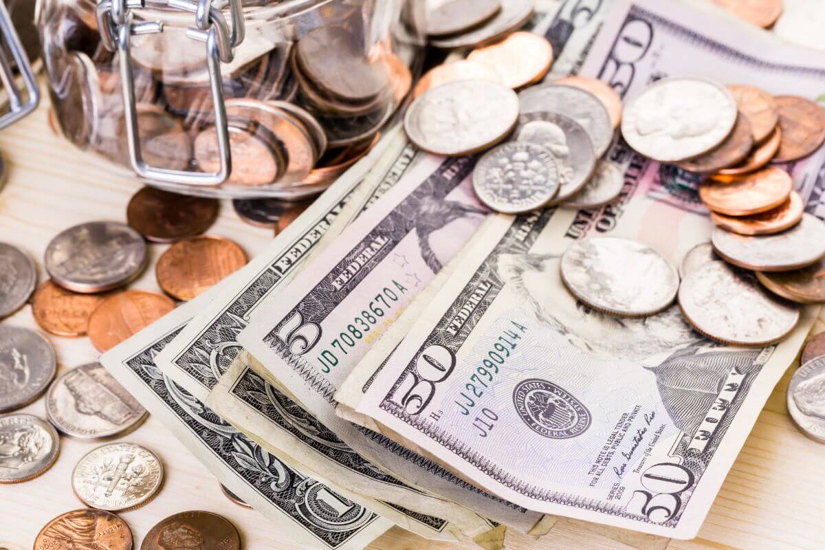 The picture shows coins on the table, dollar bills, and a jar filled with coins referring to not saving for big or future expenses that can go into debt.