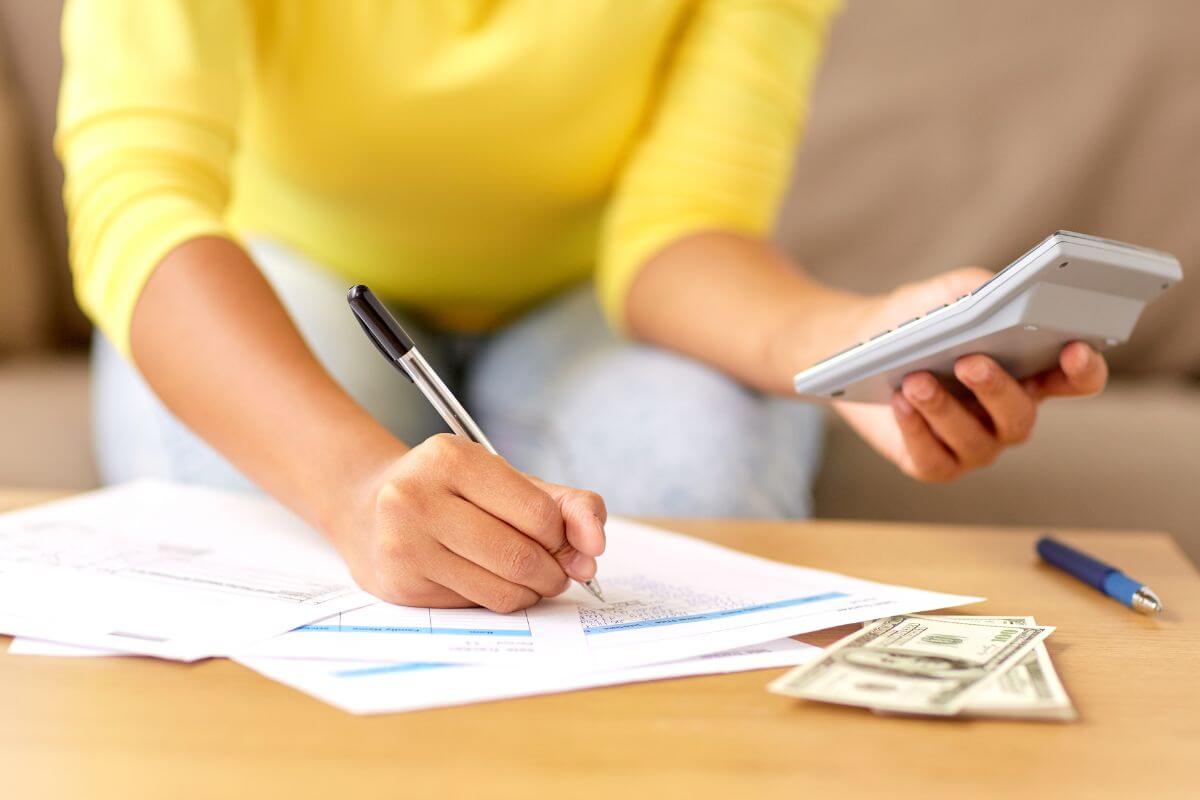 The image shows dollar bills on the table, a pen, and a girl holding a calculator while writing on the papers which means neglecting to adjust for changes in income or expenses can lead to overspending or failing short.