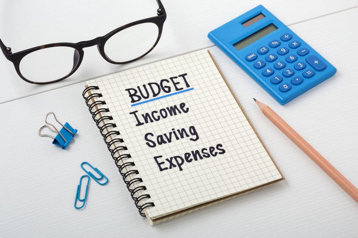 The picture of a mini calculator, clips, a pen, an eyeglass, and a notebook with notes referring to getting your budget in gear for the new year to help you reach financial freedom one step at a time.