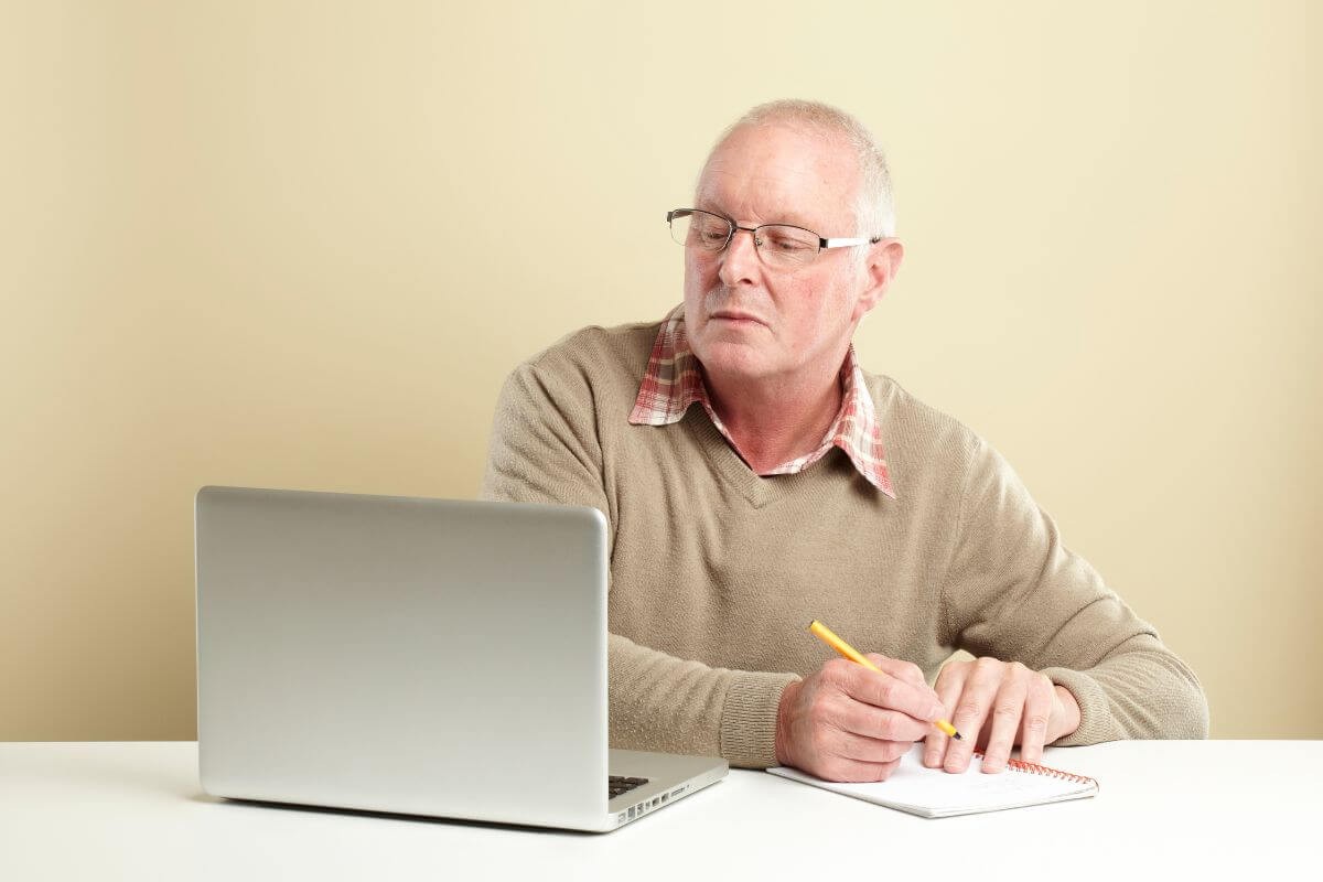 The picture tells about a senior man looking at his laptop while writing in the notebook which means that finding specific bills you get discounts on during retirement can help stretch your retirement budget even further.