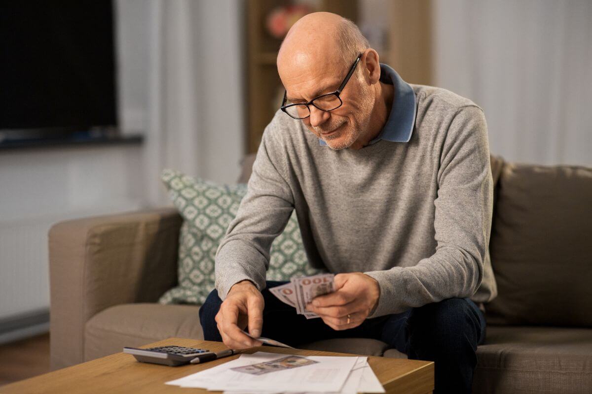 The photo shows a calculator, papers on the table, and a senior counting dollar bills, referring to increased savings each time to grow your savings without feeling the pinch.