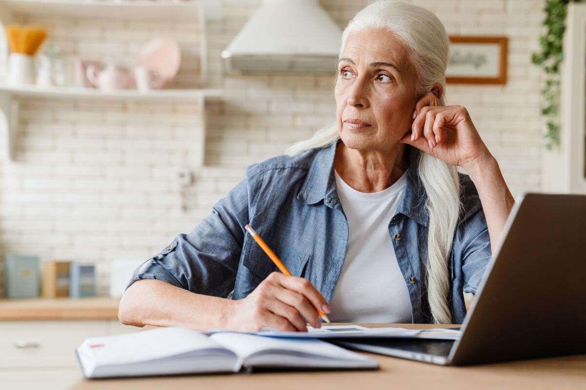 The picture shows a notebook, a laptop, and a senior writing on the papers while looking out the window thinking which refers to looking into catch-up contributions if you're 50 or older to save money than the usual limits and make up for lost time.