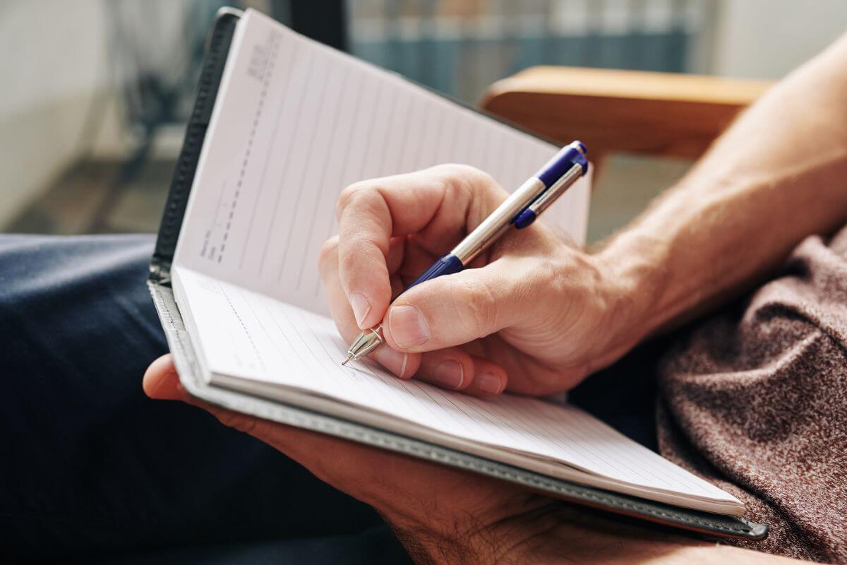 The photo tells about a man's hand and writing on a notebook which refers to writing down positive money affirmations and repeating them daily helps keep focused on achieving your money goals.
