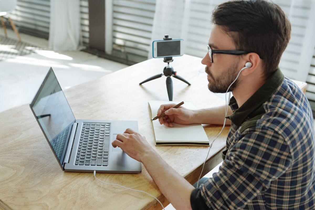 The photo shows a phone and stand, and a man writing down on a notebook while watching through a laptop indicating that educating yourself on personal finance and investments gives you the tools to grow wealth and make smarter financial choices.