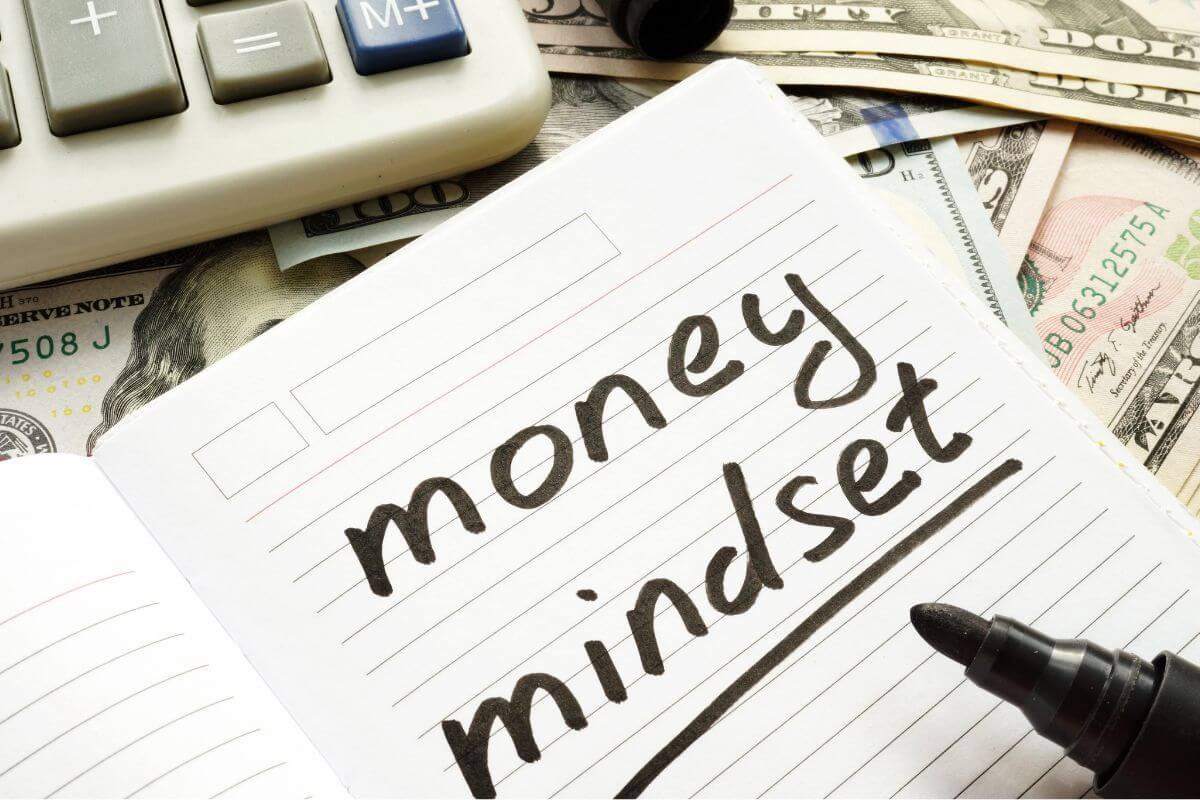 The picture shows a calculator, dollar bills, a marker, and a notebook with notes indicating that using a money mindset to build wealth can help you make decisions that lead to financial freedom.