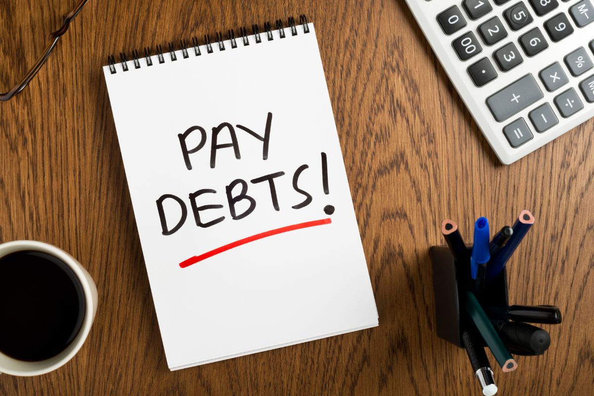 The image shows a calculator, a coffee, pens, and a piece of paper with notes referring to focus on paying off bad debts to free up more cash for savings and investments.