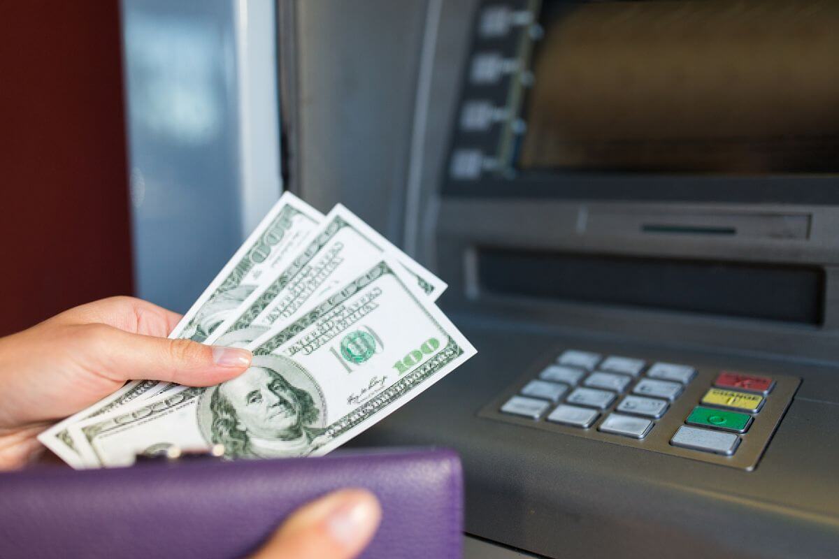 The photo tells about an Atm machine and a girl's hand putting the money inside her wallet which means to withdraw emergency cash to ensure you can buy essentials during a wildfire or hurricane evacuation.