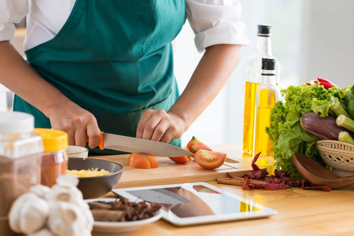 The picture describes vegetables, spices, oils, a tablet used to watch tutorials, and a man's hand cutting tomatoes, referring to how cooking meals from scratch using basic ingredients can save you hundreds of dollars each month.