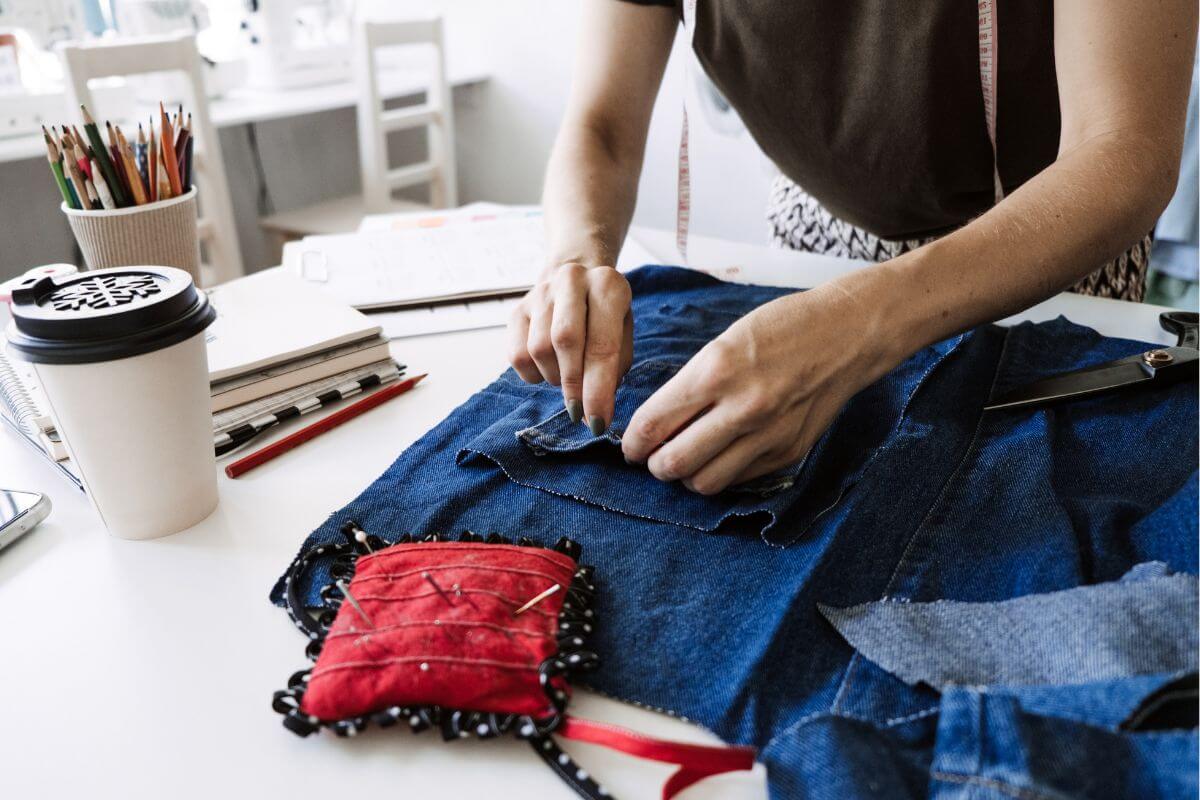 The image tells about pens, notebooks, a cup of coffee, and a girl repairing a jean indicating to sew and mend clothing to extend its life which means spending less on replacements and helping you appreciate the value of what you already own.