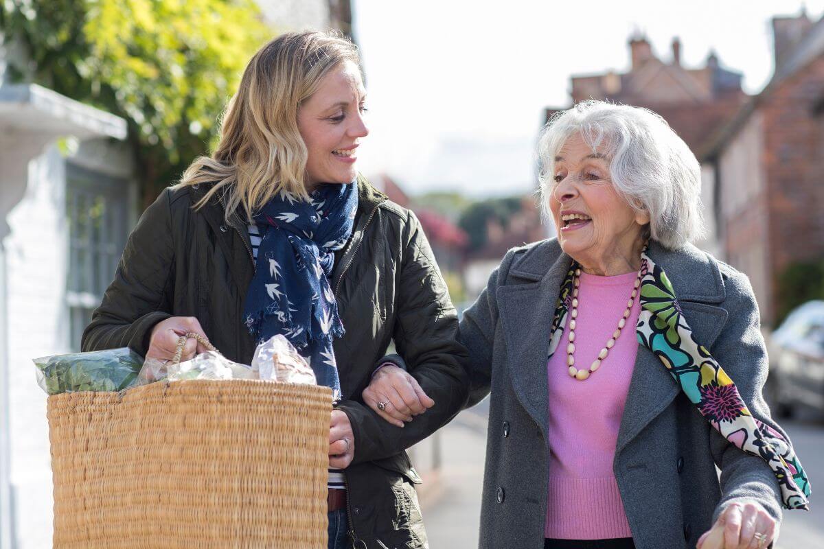 The photo shows a young girl assisting a senior with her bag full of groceries, which means practicing bartering services with neighbors, which helps keep money in your wallet and is a practical way to get things done.