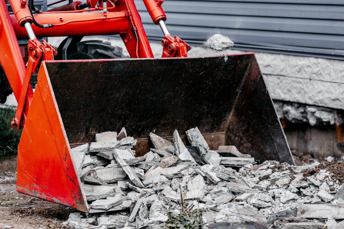 The photo tells about the bucket of a tractor collecting the broken cement which means clearing debris and mitigating the outside help protect your property from wildfire damage and give you peace of mind before evacuation orders.
