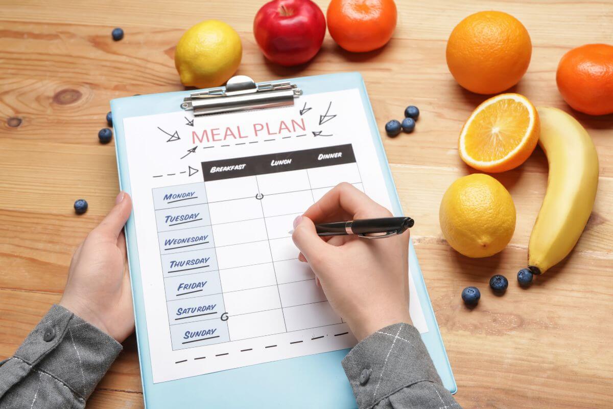 The image of fruits on the table and a girl's hand and writing on paper on a clipboard which means to plan meals carefully to reduce food waste also helps you avoid impulse purchases and makes it easier to budget for groceries.
