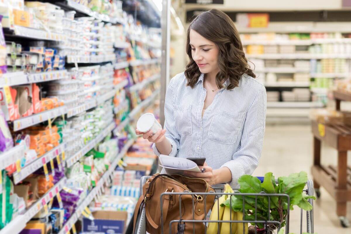 The picture tells about a cart full of fruits and vegetables, and a girl picking an item in the store referring to living within your means and prioritizing needs over wants helps you save money and build a stable future.