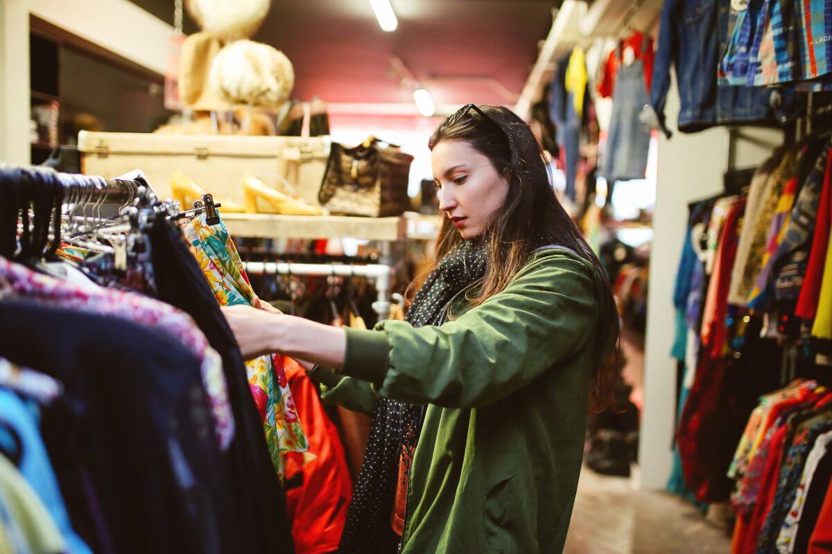 The image shows a girl picking up clothes in a store which refers to shopping secondhand for clothing that is better for your wallet and reduces waste.