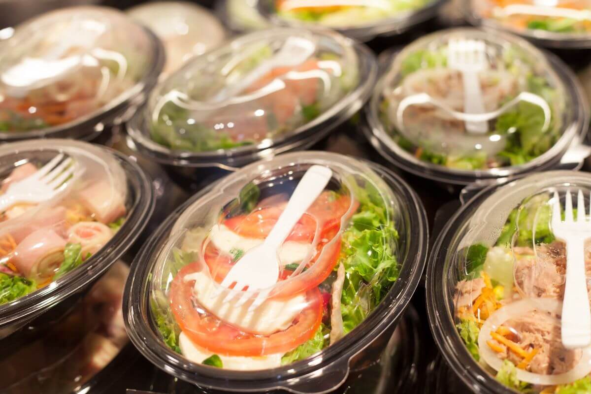 The photo tells about pre-pack salads with disposable forks which means ready-made salad kits with dressings are pricier than buying ingredients separately, making own salad helps save money.