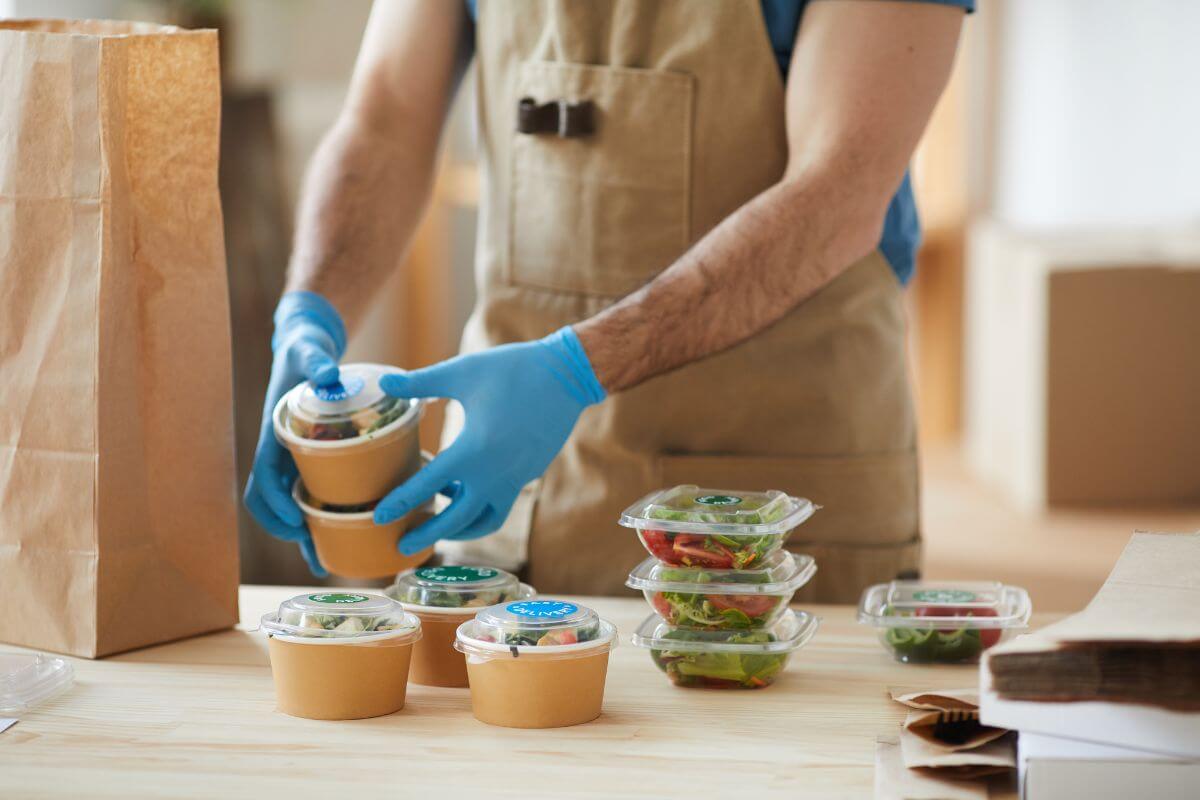 The photo tells about packed foods on the table and a man arranging them to put in a paper bag which means decorative or themed packaged foods often cost more without adding extra value, buying on store-brand versions to save money. 