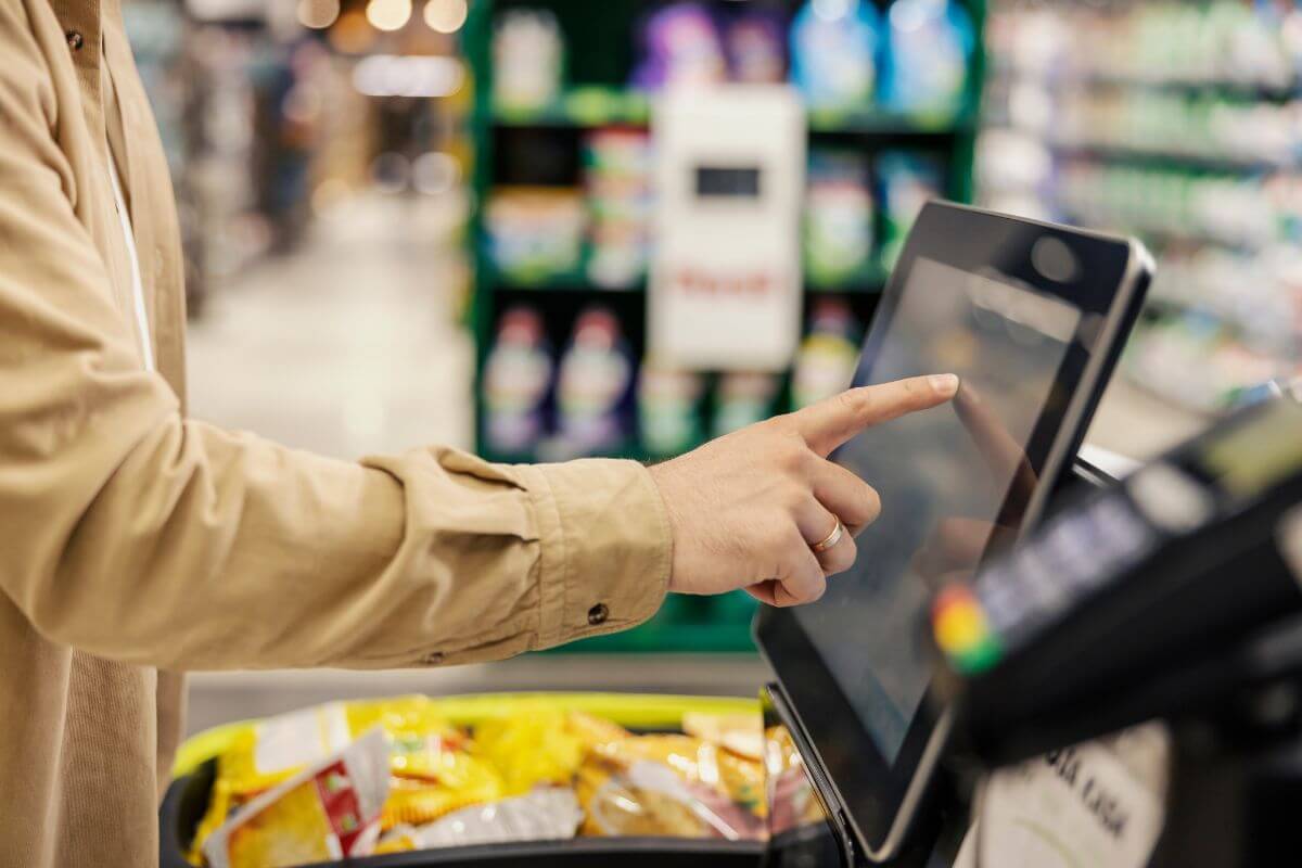 The image shows a man's hand using the machine with his cart full of items referring to impulse purchases at checkout are designed to tempt you, to skip the snacks and small items near the register to avoid overspending.