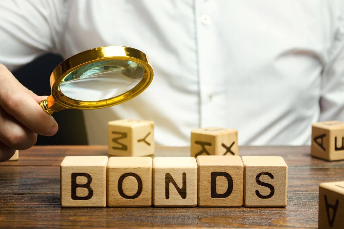 The photo shows wooden cubes with letters and a man's hand holding a magnifying glass referring to considering green bonds allow you to invest in projects focused on renewable energy infrastructure.