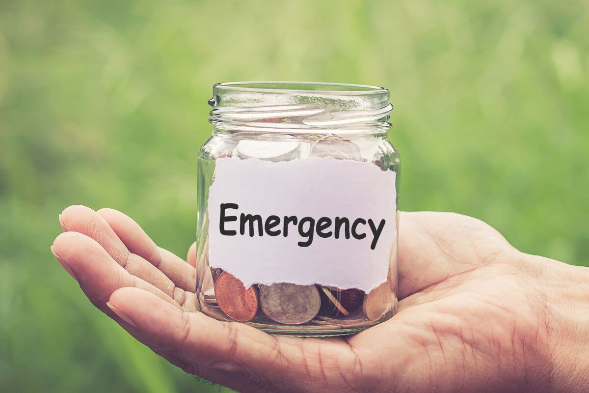 The photo shows a man's hand holding a jar with coins and with a note indicating to an alternatives to use for emergency expenses to be ready to handle surprises without going into debt.