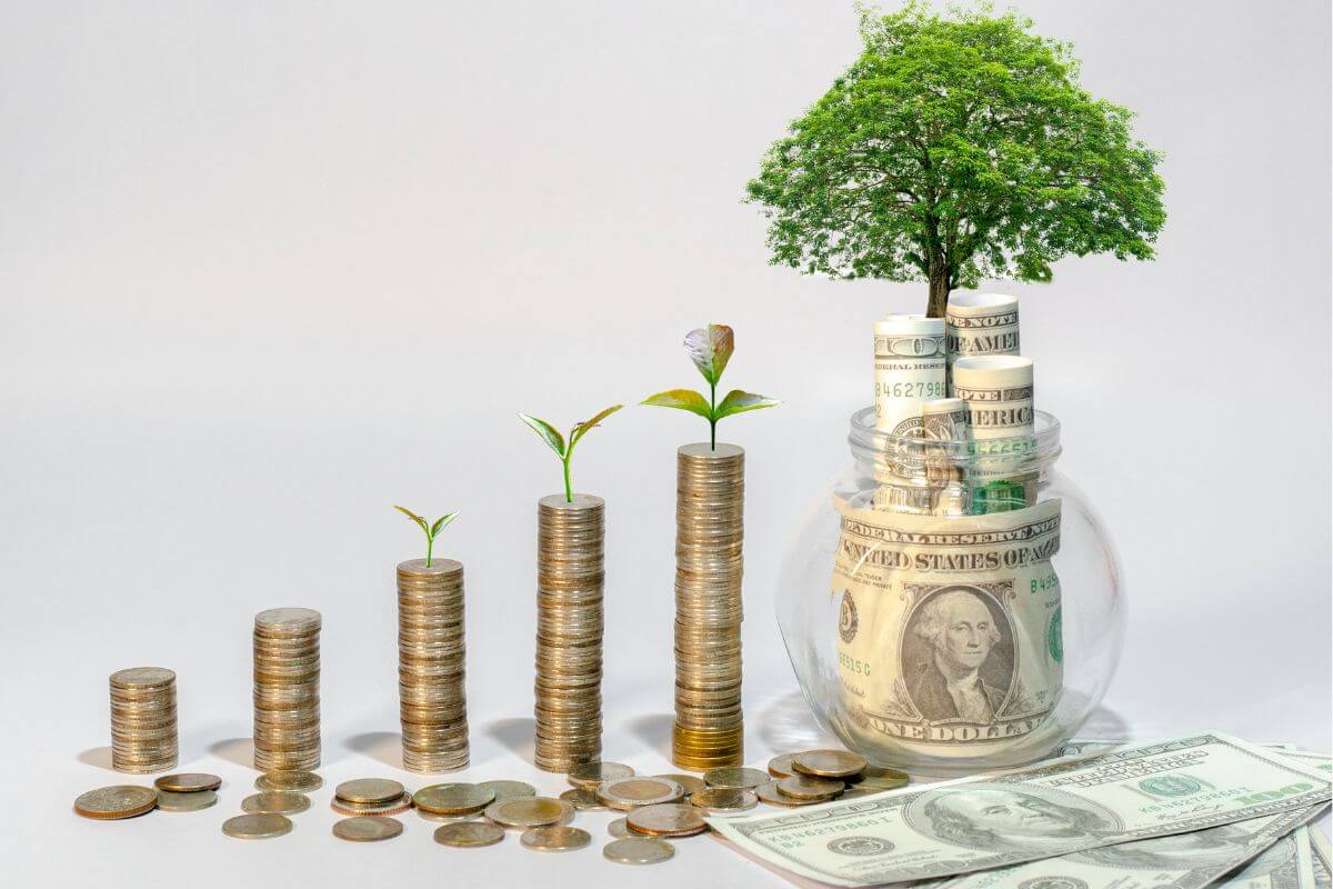 The photo tells about coin stocks, dollar bills, and a jar filled with dollar bills and a mini tree which means steps to grow savings create a financial safety net, and build a cushion for unexpected expenses.
