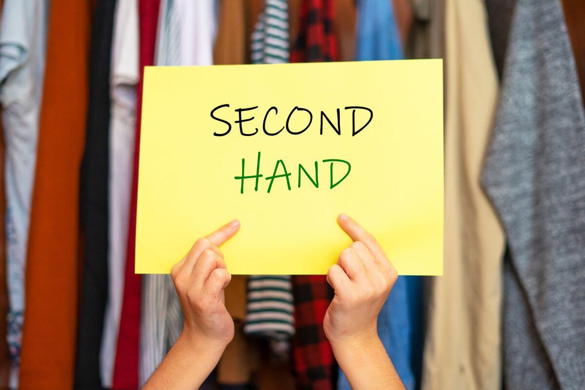 The photo of a girl's hand holding a piece of paper with notes and with clothes behind her refers to shopping secondhand for clothing to save money while reducing waste.