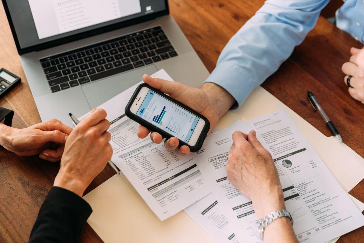 The picture tells about a laptop, papers with notes, women's hands, and a man's hand showing the screen of his phone indicating that advisors tailor financial strategies to align with their client's goals which is important when working with a financial advisor.