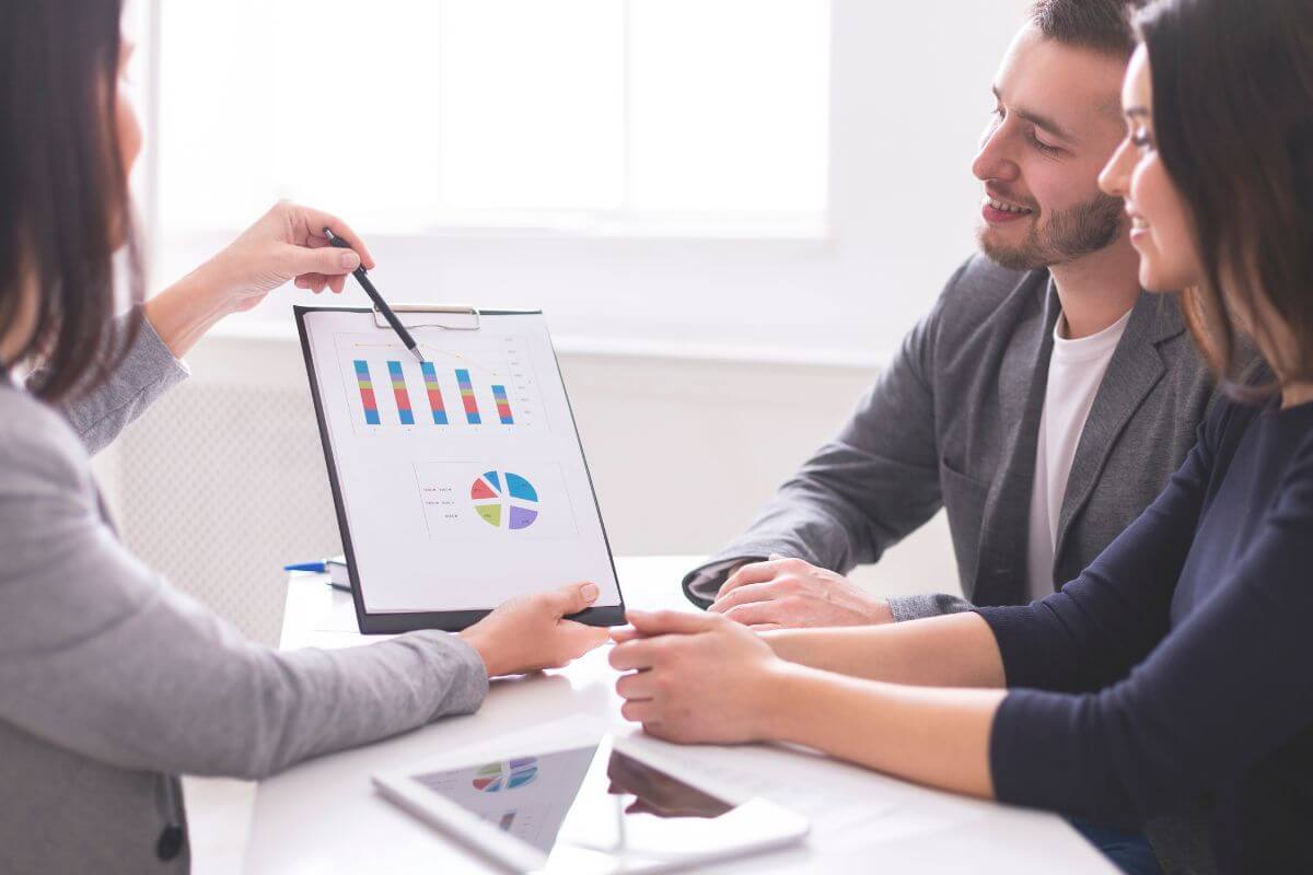 The photo tells about a tablet device on the table, a couple, and an advisor explaining the graphs on the paper which refers to often reviewing and adjusting your financial plan staying aligned with your goals, and keeping your finances on track.