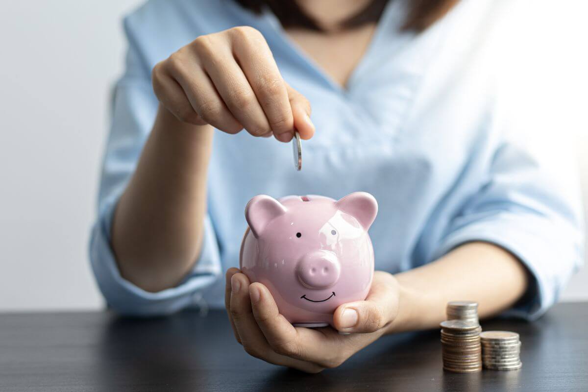 The image shows coin stock and a young girl's hand holding a piggy bank while dropping coins into it referring to how much should saved by 20's help build habits for long-term financial independence.