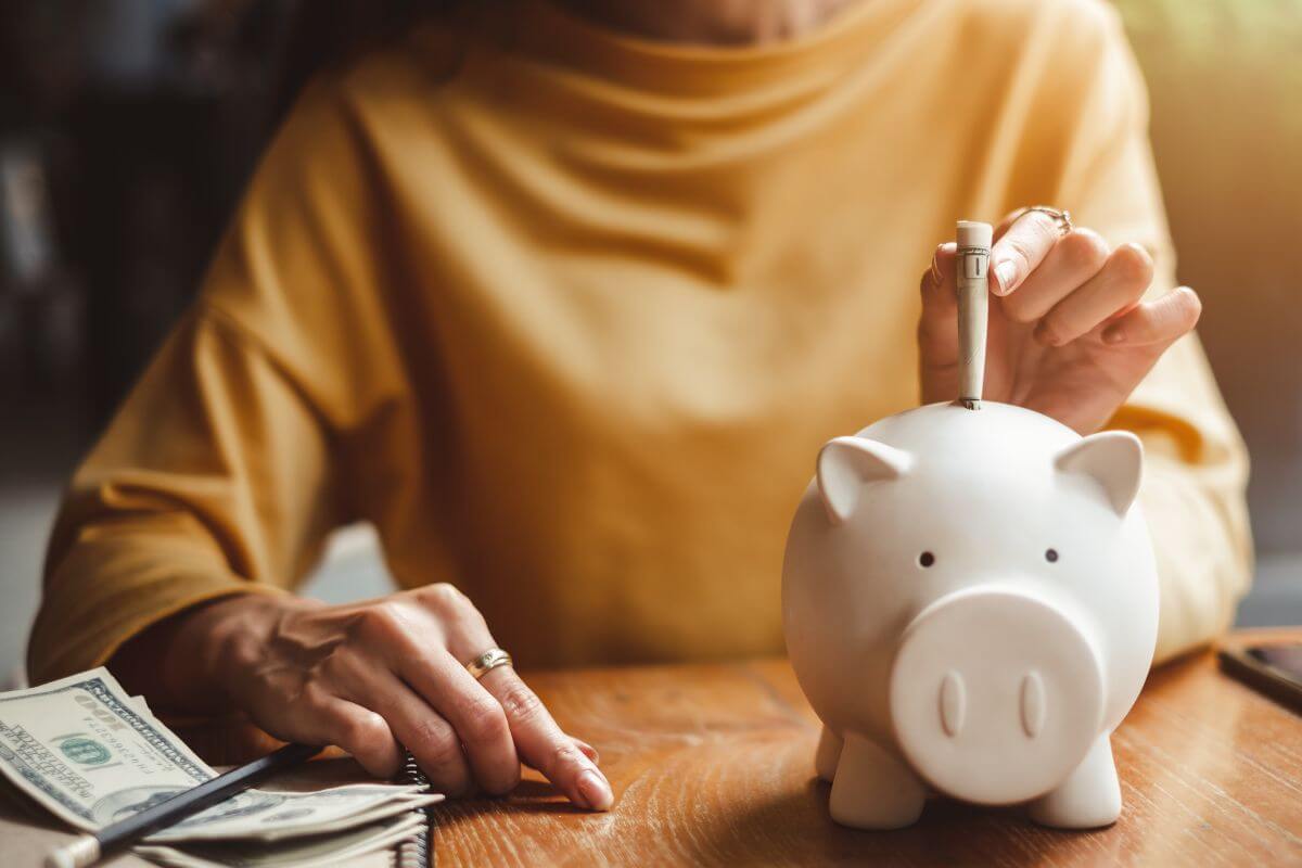 The image tells about dollar bills on the table, a notebook, a pen, and a girl putting dollar bills inside a piggy bank which means how much should save by 30, the best time to review your financial goals and adjust your savings strategy.