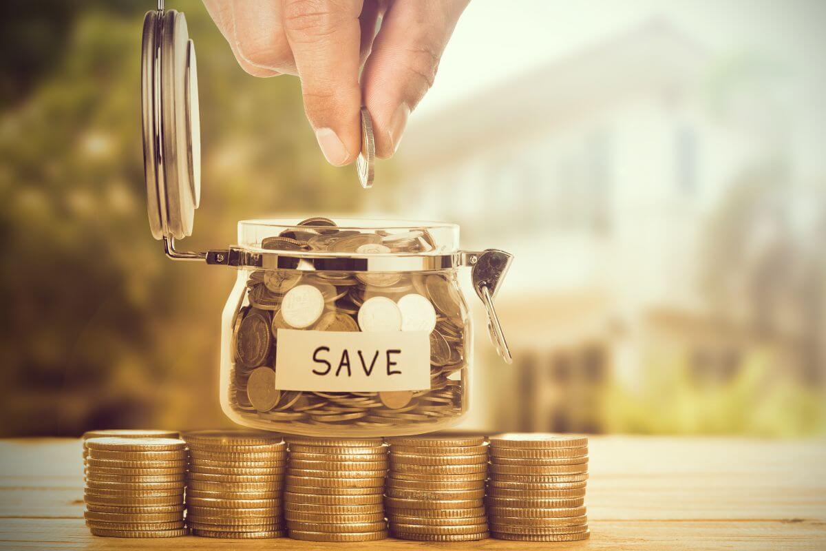 The image shows coin stocks, a jar filled with coins and notes, and a man's hand dropping a coin into it indicating how much should be saved by 40s and beyond, which is the best approach to focus on reducing debt and increasing contributions to retirement accounts.