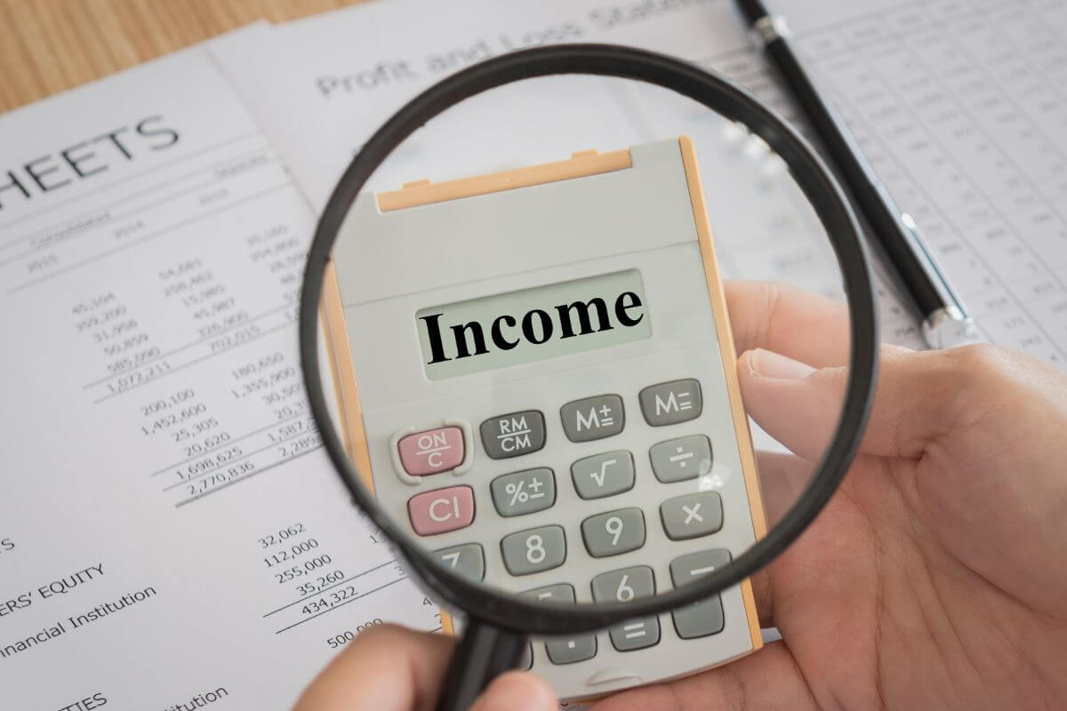 The photo of papers with notes and numbers, a pen, and a man's hand holding a calculator while using a magnifying glass which refers to the percentage of income should save each year and adjust this number based on your financial situations and future goals.
