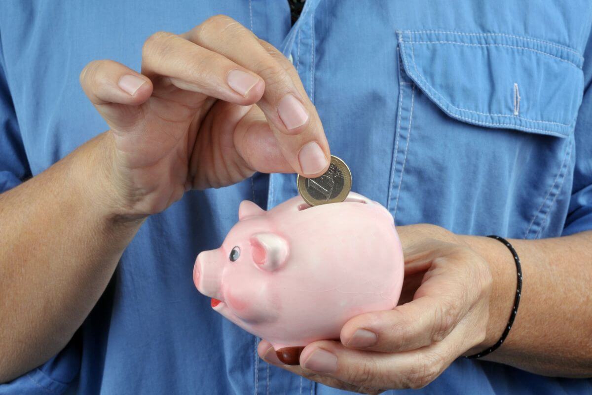 The picture shows a man's hand holding a mini piggy bank while dropping a coin into it which refers to how to adjust savings goals if started saving late, Focusing on increasing contributions helps you get closer to your goals.