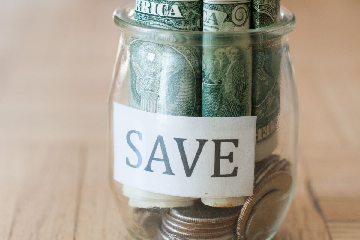 The image shows a jar filled with coins and dollar bills with a note which means that much of savings should be liquid and easily accessible to cover six months of expenses for emergencies.